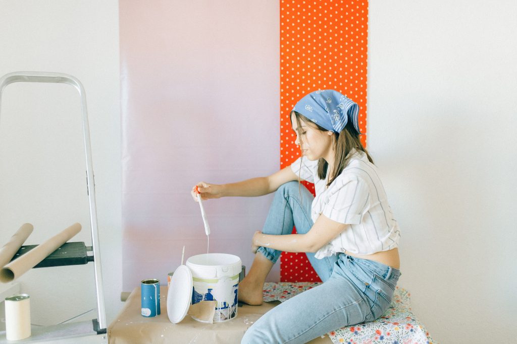 Caucasian woman sitting indoors painting with brushes and materials, wearing denim and a striped shirt.